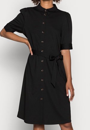 Femme portant une robe noire boutonnée longueur genou avec des manches courtes bouffantes et une ceinture en tissu nouée à la taille.