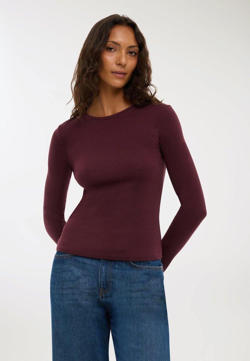 Top bordeaux a maniche lunghe realizzato in tessuto morbido con scollatura rotonda, abbinato a jeans blu a gamba larga. Silhouette dritta, dettagli minimi.