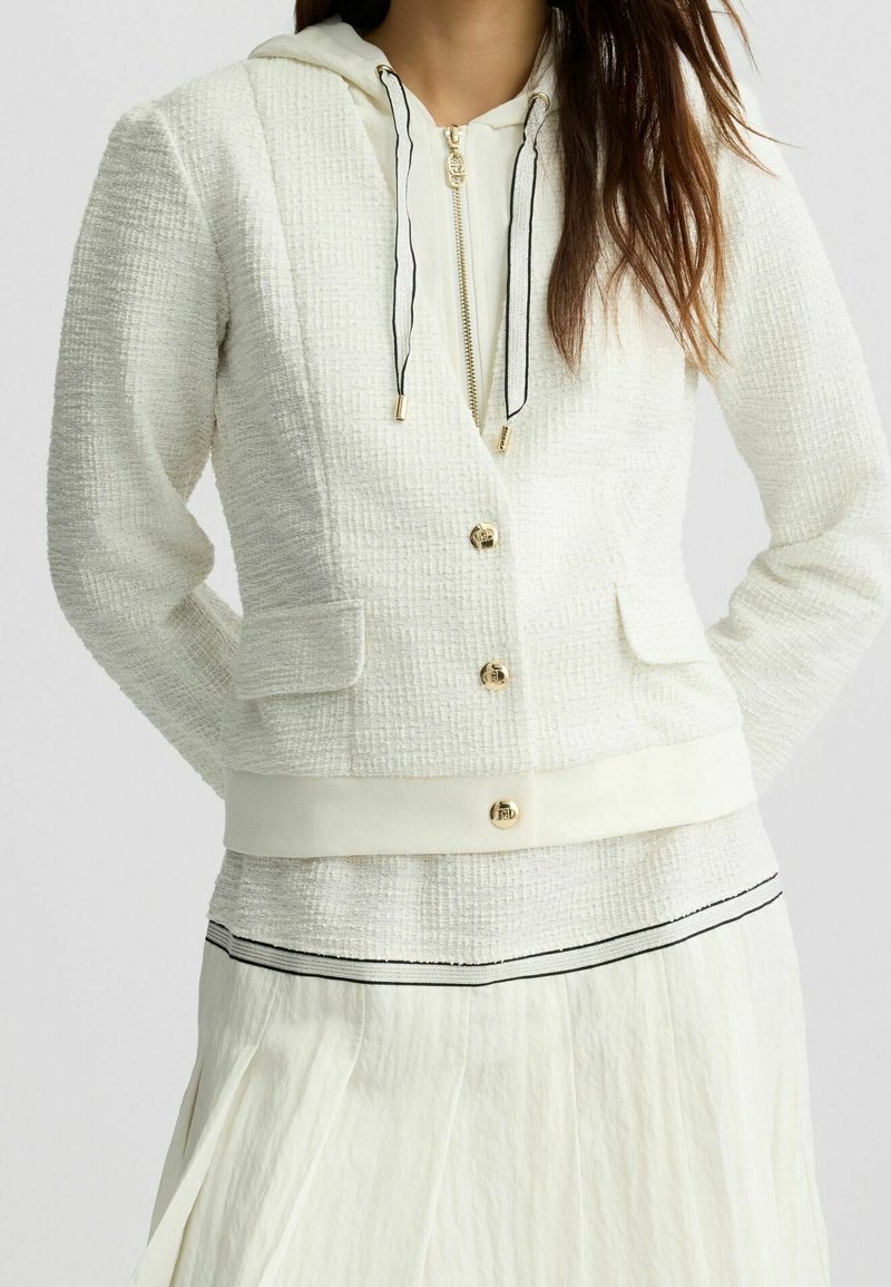 Femme portant une veste blanche texturée par-dessus un pull à capuche blanc zippé et une jupe blanche avec un bord noir à l'ourlet.