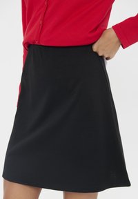 Črna A-linija krila iz mehkega materiala, z elastičnim pasom. V kombinaciji z rdečo srajco na gumbe, ki ima gladko, mat površino.