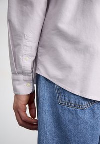 Chemise en coton lilas clair avec poignets à boutons, portée avec un jean en denim bleu. Présente un ourlet arrondi et une texture subtile.