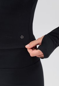 Černý atletický top s dlouhým rukávem a přiléhavým střihem. Má bezešvý pas a nenápadné logo na boku. Měkká elastická látka.