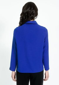 Kék színű, hosszú ujjú blúz, sima anyagból, ívelt aljjal. Gombos mandzsettákkal rendelkezik, és letisztult, egyszerű kialakítással bír.