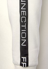 Weiße Jogginghose mit einem schwarzen vertikalen Streifen und weißem Text "CONNECTION" und "F" an der Seite. Weicher, strukturiert Stoff.