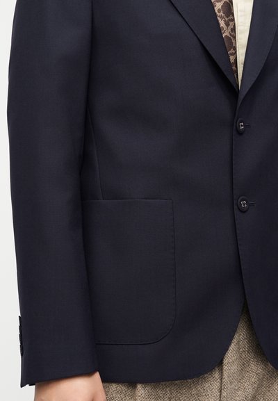 Σκούρο μπλε σακάκι με μπροστινή τσέπη, κουμπωμένο με δυο κουμπιά, φορεμένο πάνω από γραβάτα με σχέδιο και ανοιχτό πουκάμισο, συνδυασμένο με μπεζ υφασμένα παντελόνια.
