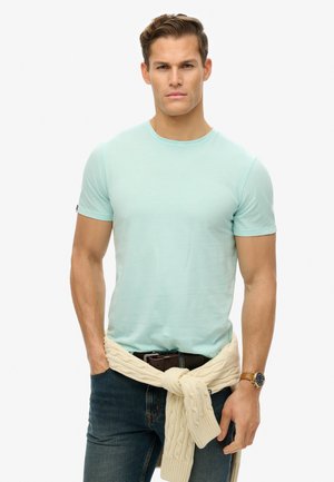 Camiseta de manga corta verde menta, con cuello redondo, llevada con jeans azules y un suéter de punto color crema atado en la cintura. Reloj marrón en la muñeca.