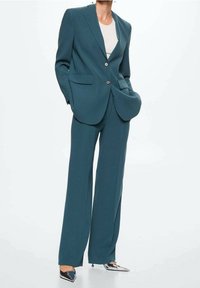 Costume sarcelle comprenant une veste ajustée avec deux boutons et un pantalon à jambes larges. Tissu lisse, design minimaliste et col pointu. Talons argentés.