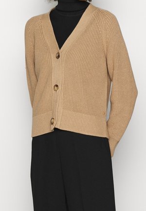 Beige gebreid vest met v-hals, voorzien van vier bruine knopen, geribbelde zoom en lange mouwen, gedragen over een zwarte coltrui.