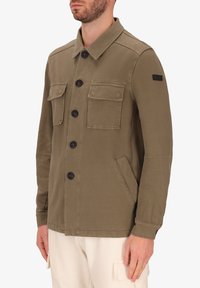 Giacca verde oliva realizzata in misto cotone, con chiusura a bottoni, due tasche sul petto, tasche laterali e una patch nera con logo sulla manica.