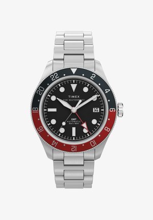 Montre Timex Waterbury GMT en acier inoxydable avec cadran noir, lunette 24 heures rouge et noire, aiguilles argentées, fenêtre de date et bracelet à maillons métalliques.