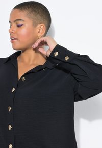 Una mujer con el cabello corto mira ligeramente hacia la izquierda, vestida con una camisa negra decorada con botones dorados, con la mano levantada cerca de su cuello, frente a un fondo blanco.