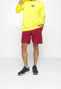 Camiseta deportiva de manga larga de color amarillo brillante con logo negro, combinada con pantalones cortos color burdeos con un detalle negro, y zapatillas de correr negras.