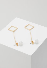 Goldene geometrische Ohrhängern mit quadratischen Formen, einem Perlenakzent und einer dünnen Metallkette, vor einem hellgrauen Hintergrund.