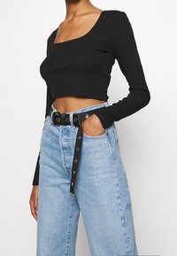 Nainen, jolla on päällään musta pitkähihaisen crop-top, korkeavyötäröiset vaaleansiniset farkut mustalla vyöllä, käsi taskussa valkoista taustaa vasten.