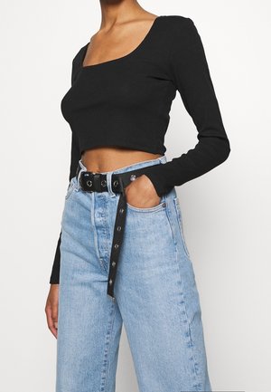 Femme portant un haut court à manches longues noir et un jean taille haute bleu clair avec une ceinture noire, main dans la poche, sur fond blanc.