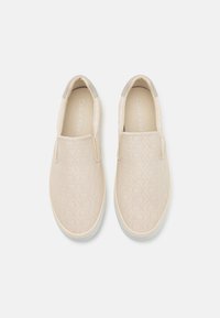 Beige slip-on sko med preget logo-mønster, gr�å detaljer ved hælen, hvit gummisåle og glatt tekstiloverdel. Klassisk uformell design.
