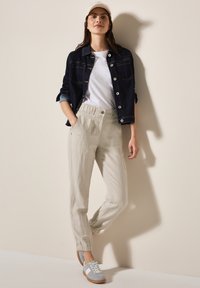 Lätta, beige avsmalnande byxor med elastiska mudd, parat med en marinblå denimjacka och en vit T-shirt. Grå sneakers fulländar outfiten.