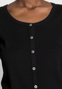 Svart stickad cardigan med ribbad halsringning, klassisk knäppning fram med rund, iriserande knappar och en figurnära passform.