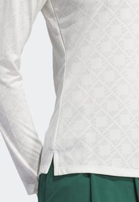 Bílé sportovní tričko s dlouhým rukávem, strukturovaným vzorem a bočními rozparky, doplněné zelenými šortkami. Vyrobené z prodyšné látky.