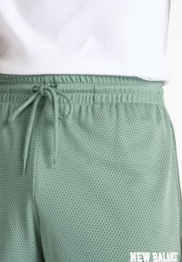 Lys grønne mesh-shorts med elastisk talje, snøre og "NEW BALANCE"-branding nederst til højre.