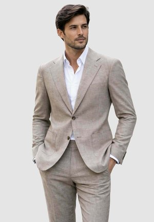 Uomo con abito beige e camicia bianca, mani in tasca, guarda di lato su sfondo chiaro e uniforme.