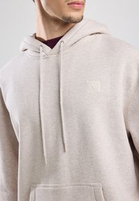 Beige Kapuzenpullover aus weichem Stoff, mit einer Fronttasche, Kordeln und einem kleinen strukturierten Logo auf der Brust.