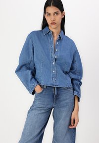 Camicia di jeans blu chiaro con bottoni, dal taglio morbido, caratterizzata da colletto, maniche over e tasche frontali. Abbinata a jeans abbinati.