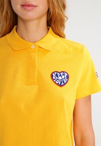 Żółta koszulka polo z bawełny, z kołnierzykiem, jednym guzikiem oraz czerwoną łatą w kształcie serca z napisem "KEEP LOVING" w niebieskim kolorze.