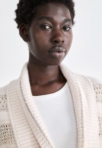 Helle beige gestrickte Strickjacke mit einem strukturierten Muster und glänzenden silbernen Verzierungen, getragen über einem weißen gerippten Shirt.