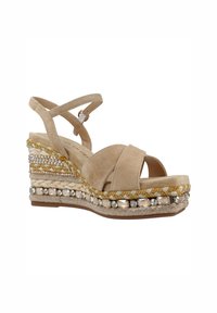 Sandale compensée en suede beige avec des brides entrecroisées, une semelle intermédiaire tressée, et ornée de perles et de paillettes pour une texture et des détails.