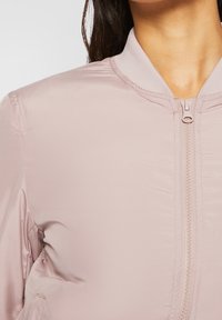 Primo piano di una persona che indossa una giacca a zip rosa chiaro con colletto a coste e lunghi capelli scuri visibili sul lato sinistro.