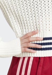 Suéter branco de malha com bainha canelada, apresentando riscas azul-marinho. Mão posicionada na cintura do suéter, com calças vermelhas a mostrar riscas cor-de-rosa.