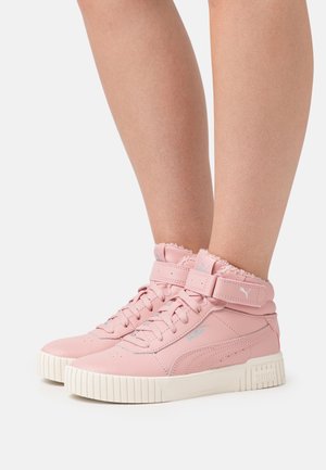 Baskets montantes roses avec semelles blanches, lacets et bandes Velcro, portées sur des jambes nues sur un fond clair uni.