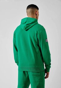 Sweat à capuche vert en tissu doux avec une capuche, ourlet et poignets côtelés, présentant un patch logo sur la manche gauche. Porté avec un pantalon assorti.