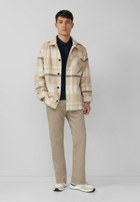 Beżowa kratkowana kurtka z ciemnymi guzikami, noszona na granatowym polo, w zestawieniu z jasnobeżowymi spodniami i białymi trampkami z zielonymi akcentami.