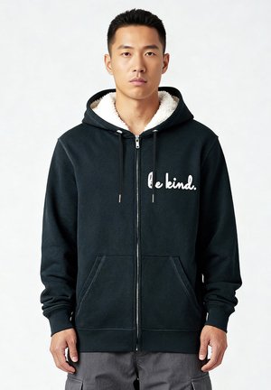 Schwarze Zip-Hoodie mit einem kuscheligen Innenfutter, ausgestattet mit dem Schriftzug "sei freundlich." in weißem Text, zwei vorderen Taschen und einer Kapuze mit Kordelzug.
