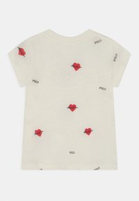 Krátký rukáv, krémové tričko s rozptýlenou červenou výšivkou srdcí a nápisem „POLO“. Bavlněný materiál, volný střih, bez výrazné textury.