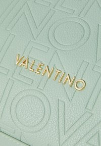 Mėtų žalios tekstūruotos odos krepšys, pasižymintis įspaustais užrašais ir auksiniu "VALENTINO" logotipu kaip išskirtiniu akcentu.