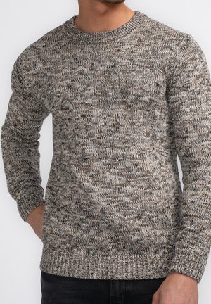 Mies, joka on pukeutunut beigeen ja ruskeaan kirjoneulepaitaan pitkähihaisena, seisoo käsi taskussa ja katsoo hieman vasemmalle.