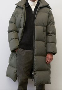 Person wearing a long olive green puffer coat over a black zip sweater and white turtleneck, standing against a plain light background.