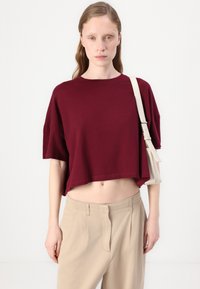 Burgundské zkrácené tričko s krátkým rukávem, vyrobené z bavlny. V páru s béžovými plisovanými kalhotami. Model drží světlou tašku.