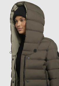 Aizpildīta olīvzaļā jaka ar lielu, salokāmu kapuci, ar rāvējslēdzēju un pogu aizdari, un gludu, matētu tekstūru. Melna adīta cepure zem tās.