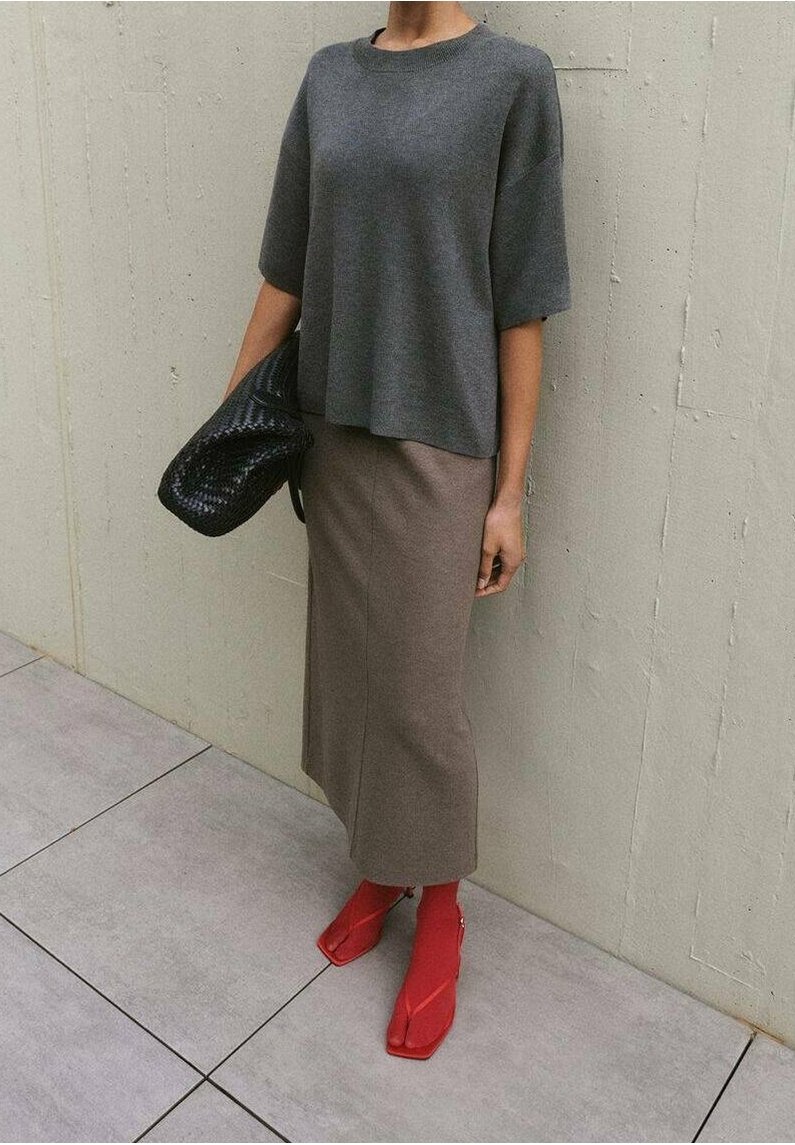 Personne portant un haut gris foncé à manches courtes, une longue jupe taupe, des chaussures rouges à bouts ouverts et tenant une pochette noire texturée contre un mur beige.