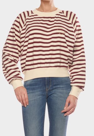 Person trägt einen cremefarbenen und burgunderroten gestreiften Pullover mit langen Ärmeln und elastischen Bündchen sowie eine blaue Jeans und steht vor einem schlichten Hintergrund.