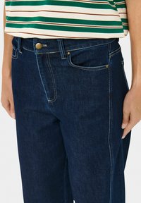 Isik, kellel on seljas tume sinine teksapüksid ja roheline-valge triibuline särk, käed lõdvestatult külgedel.