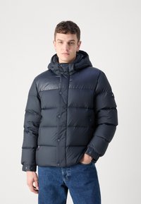 Giubbotto imbottito di colore blu navy scuro con cappuccio, caratterizzato da un design trapuntato, bottoni a pressione e tasche laterali. Realizzato in materiale lucido e isolante.