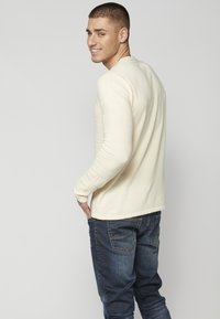 Helles beiges Langarmshirt mit geripptem Textur an den Ärmeln, kombiniert mit dunkelblauen Jeans, von hinten gezeigt. Slim-Fit-Design.