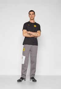 Sort kortærmet Puma t-shirt med gult logo, parret med grå sweatpants med hvide sidestykker og logo, afsluttet med sorte sneakers.