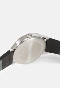 Bracelet en acier inoxydable avec un dos rond, un bracelet en cuir noir et le nom de la marque gravé. Design résistant à l'eau avec des détails texturés.