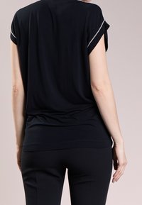 Persona con una camiseta negra de manga corta con ribetes plateados en las mangas y pantalones negros, de pie frente a un fondo neutro, vista por detrás.